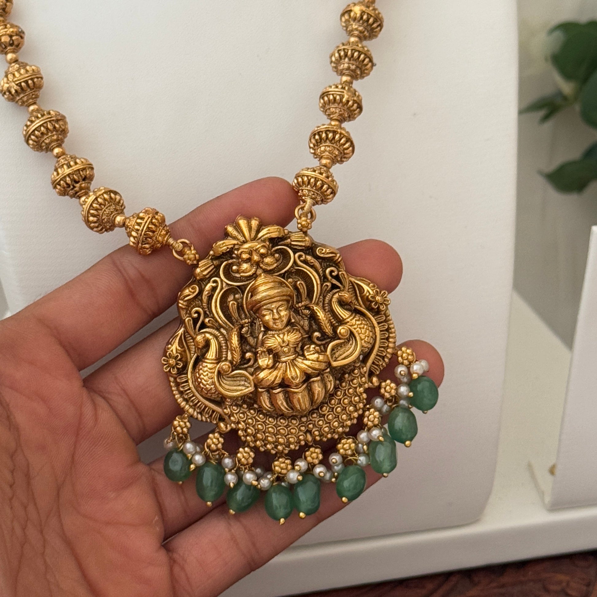 Gold necklace with a large decorative pendant held by a hand against a neutral background