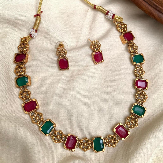 Gold necklace with red and green gemstones and matching earrings on a beige fabric background